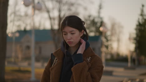 a girl wearing a brown coat and a hoodie is walking alone in a park, looking visibly unhappy and lost in thought. the soft, warm light of the golden hour enhances the somber mood