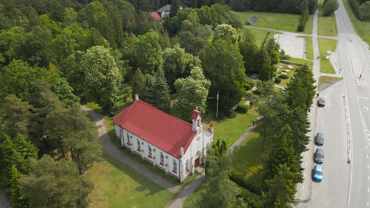 Gorgeous aerial revealing footage of an old and historic medieval white church in the sunny countryside by a road in europe during day time. Footage flies backwards and reveals large forest around it.