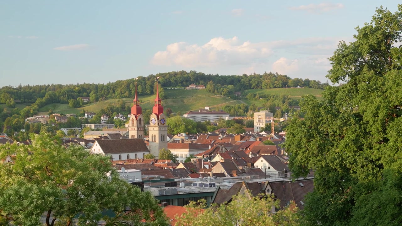 Vista panoramica di una cittadina europea immersa tra dolci colline verdi. La scena è avvolta dalla luce calda e soffusa tipica del tardo pomeriggio o del primo mattino