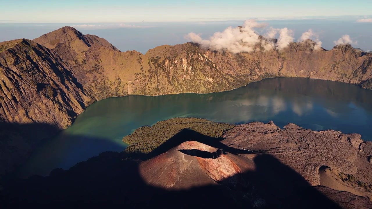 Cono vulcanico accanto a un lago di caldera sotto il bordo della montagna nella chiara luce del mattino, panoramica aerea con acqua turchese, Rinjani, Indonesia