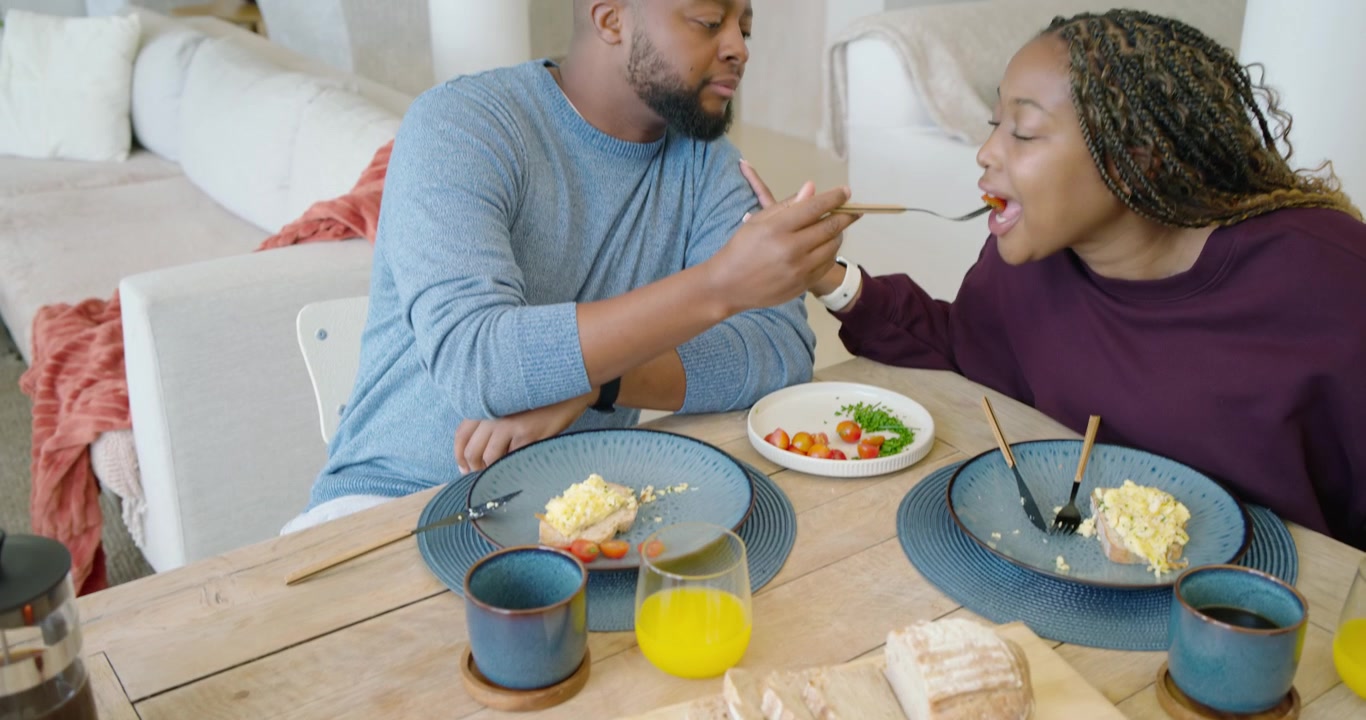 African American couple sharing breakfast at wooden table, woman touching shoulder, man feeding her