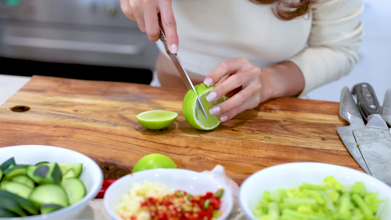 A person slices a lime on a wooden board surrounded by fresh salad ingredients in a bright kitchen
