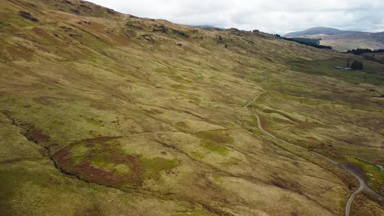 Aerial drone footage slowly gliding above a small mud rural road that is twisting and turning between scottish highlands grassy brown and yellow hills during a cloudy day while mountains are visible.
