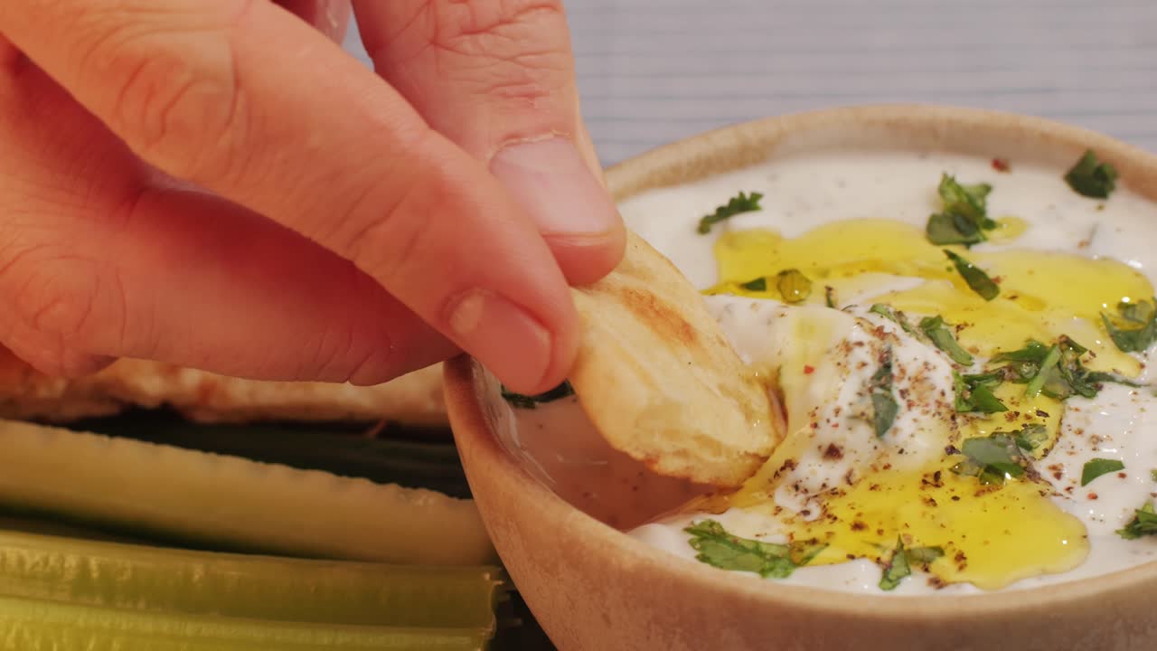 Hand dipping pita bread into a creamy yogurt dip with herbs and olive oil