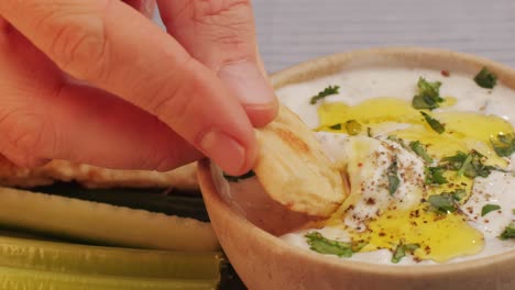 hand dipping pita bread into a creamy yogurt dip with herbs and olive oil