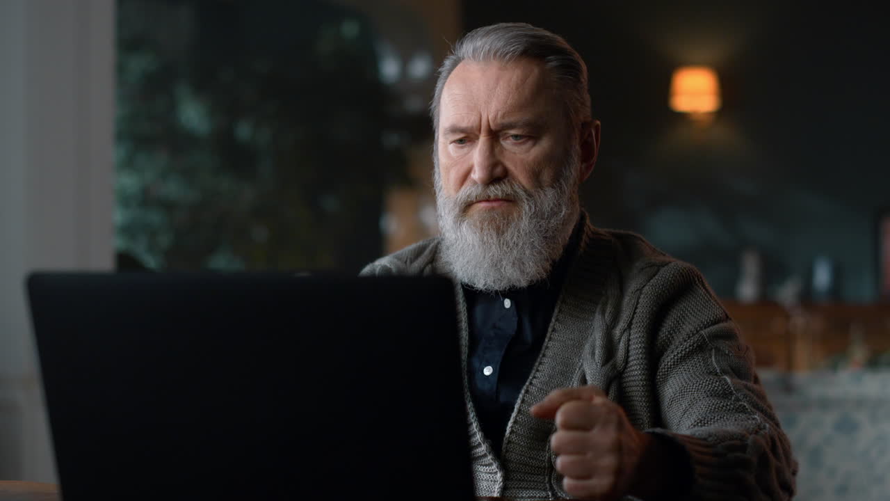 hombre de edad avanzada estricto haciendo una llamada de video en la computadora en la sala de estar de lujo