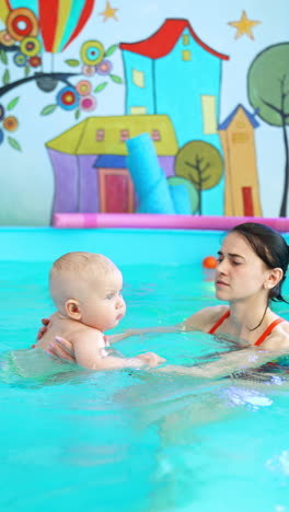 female coach teaches the toddler baby to swim in the swimming pool. calm child learning to hold in the water. vertical video.