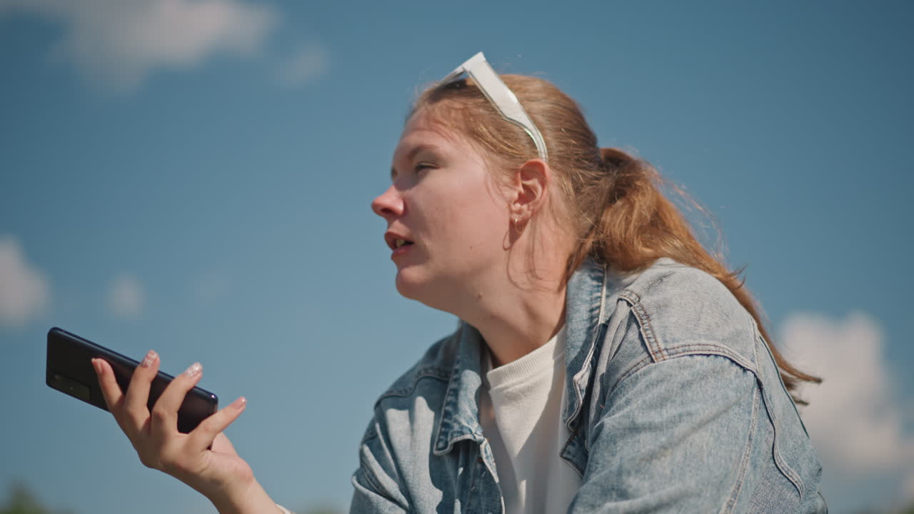 Ung hvid kvinde optager stemmebesked, denimjakke og hvidt hårbånd, håndholdt smartphone tæt på mund, udtryksfuldt frustreret ansigt, solrig blå himmel med spredte skyer, udendørs parkmiljø.