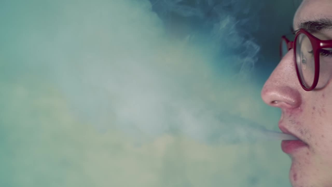 Young pimply man in red glasses vaping and exhales smoke indoor. Side view with a cinematic move from right to left side. Close up. Slow motion