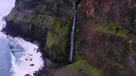 Filmische-Luftaufnahme-Des-Wasserfalls,-Der-Die-überwucherten-Berge-Hinunter-Ins-Ufer-Des-Ozeans-Auf-Der-Insel-Madeira-Fällt