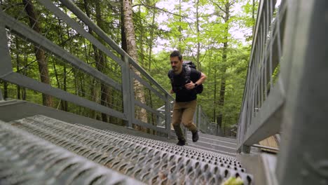 male hiker with heavy backpack quickly ascends steep metal staircase in lush green forest landscape