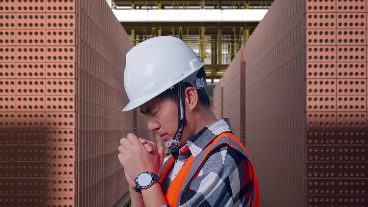 Close Up Side View Of Asian Male Engineer With Safety Helmet Pray For Something While Standing With Red Brick Packed in Stacks Are Stored