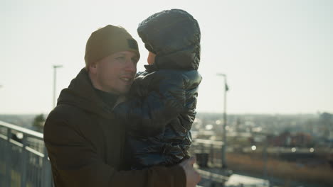 un padre está de pie cerca de una barandilla de metal, llevando a su hijo pequeño cerca de su pecho, ambos miran hacia un paisaje urbano en la distancia, ambos con chaquetas cálidas y gorras
