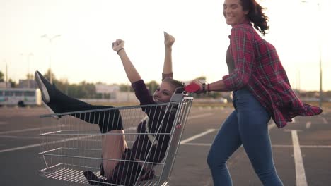 side view of a happy young woman pushing a grocery cart with her girlfriend inside in the parking by the shopping mall during