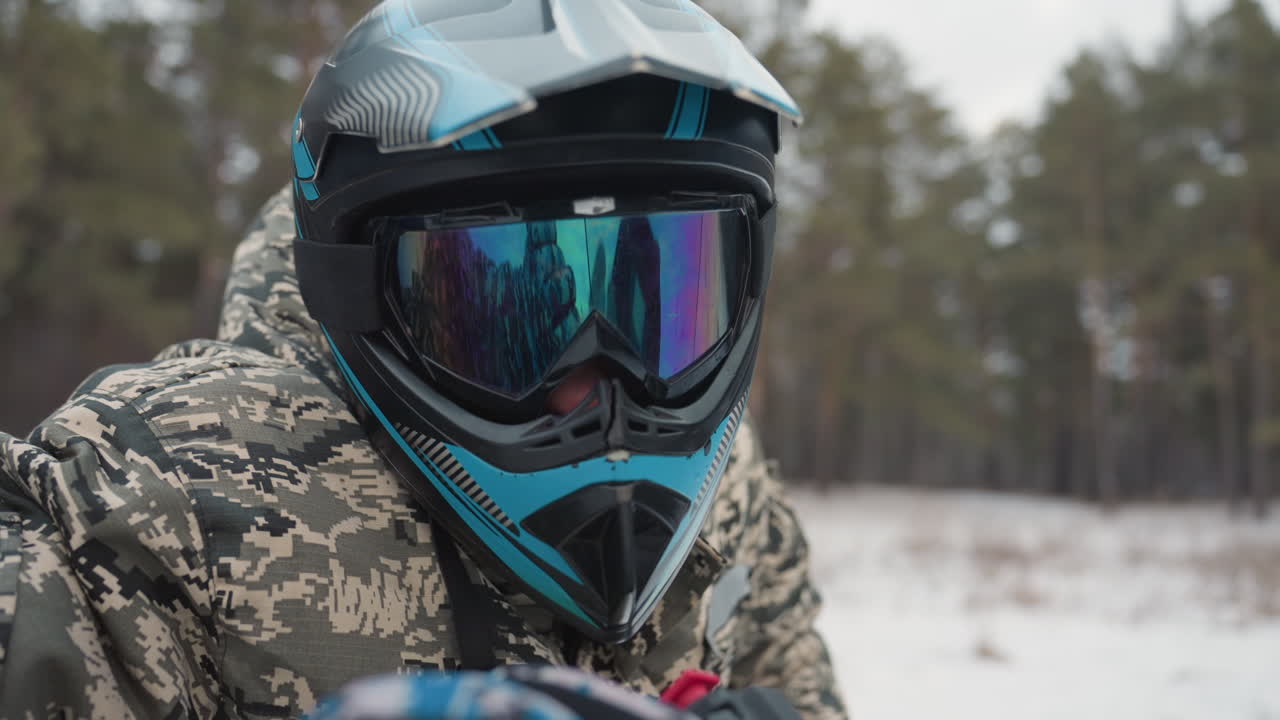 Fører på ATV i snødekt skog, kledd i kamuflasjejakke og blå hjelm med speilglassbriller, griper styret bak et klart frontrute. Speideren sjekker løypa. Furutrær i kaldt vinterlys, med diskret pust synlig.