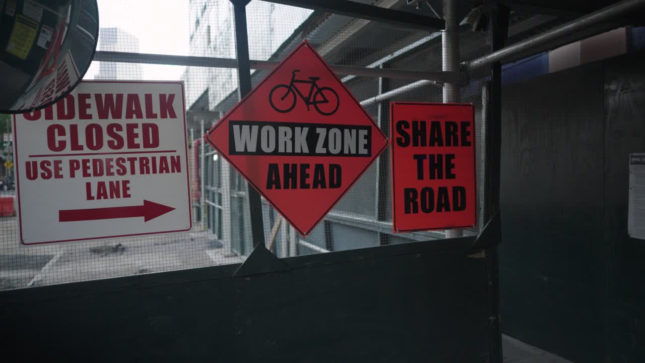 en una pasarela de andamios de construcción, señales advierten de una acera cerrada y la próxima zona de trabajo, instando al uso compartido de la carretera