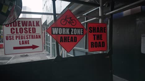 en una pasarela de andamios de construcción, señales advierten de una acera cerrada y la próxima zona de trabajo, instando al uso compartido de la carretera