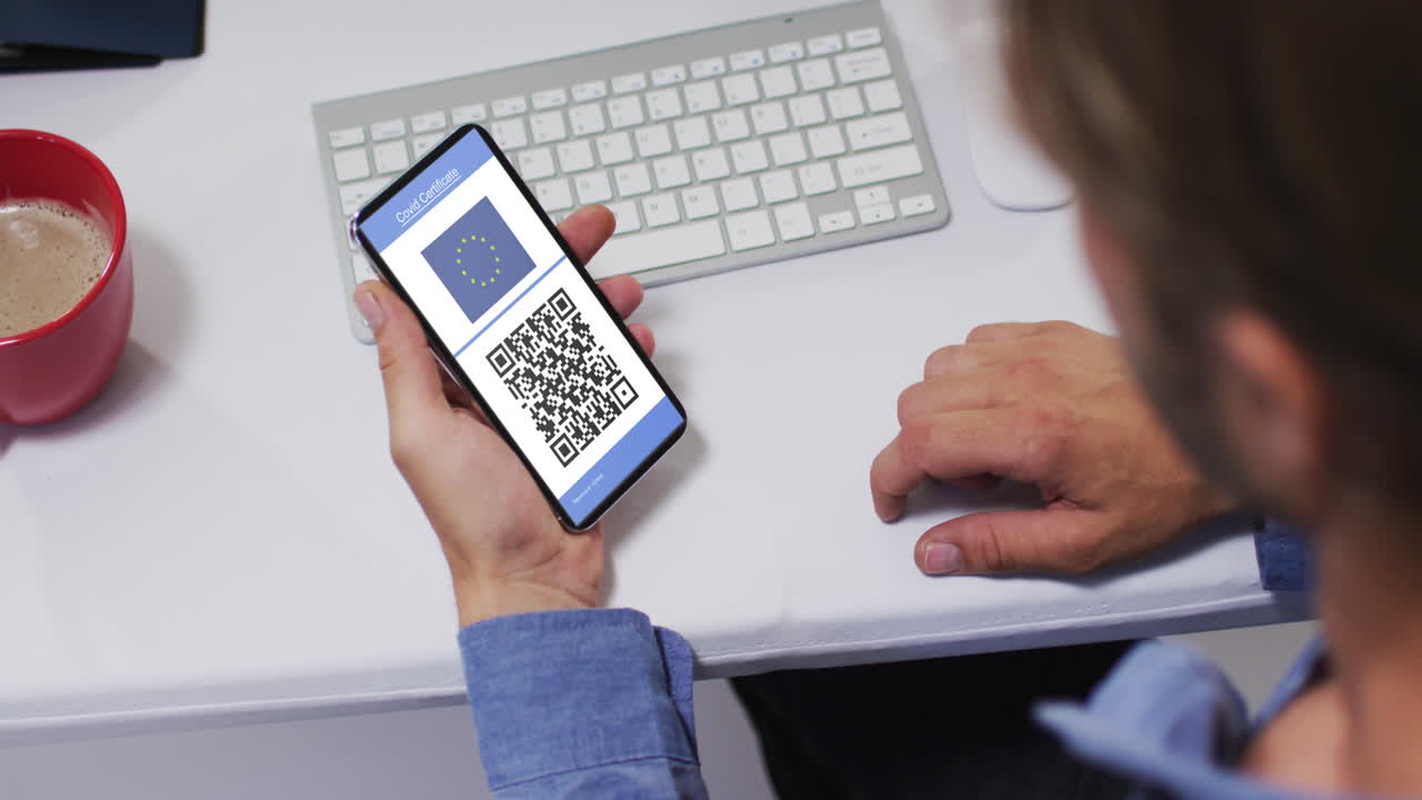 Man at desk holding smartphone with covid vaccination certificate, eu flag and qr code on screen