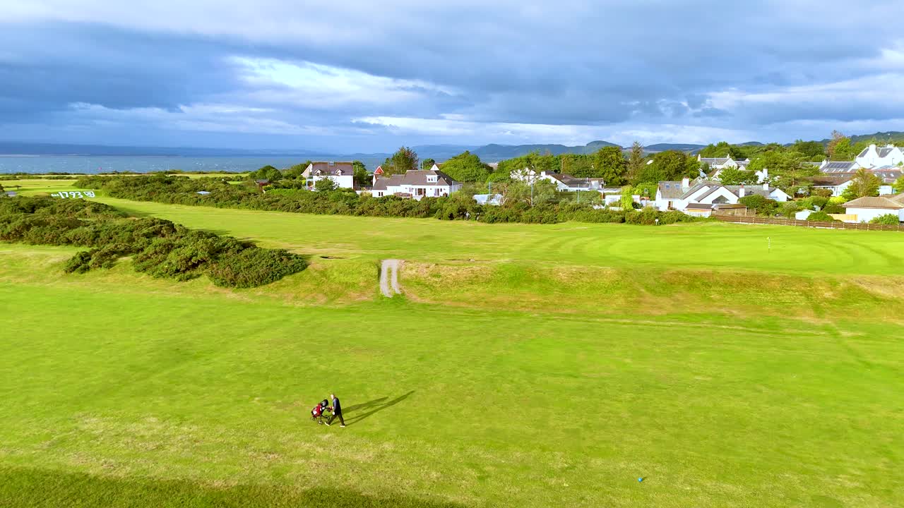 Two golfers with a caddy walk across a wide, green fairway in Rosemarkie, Scotland, under dramatic skies. Aerial view, steady camera, natural light