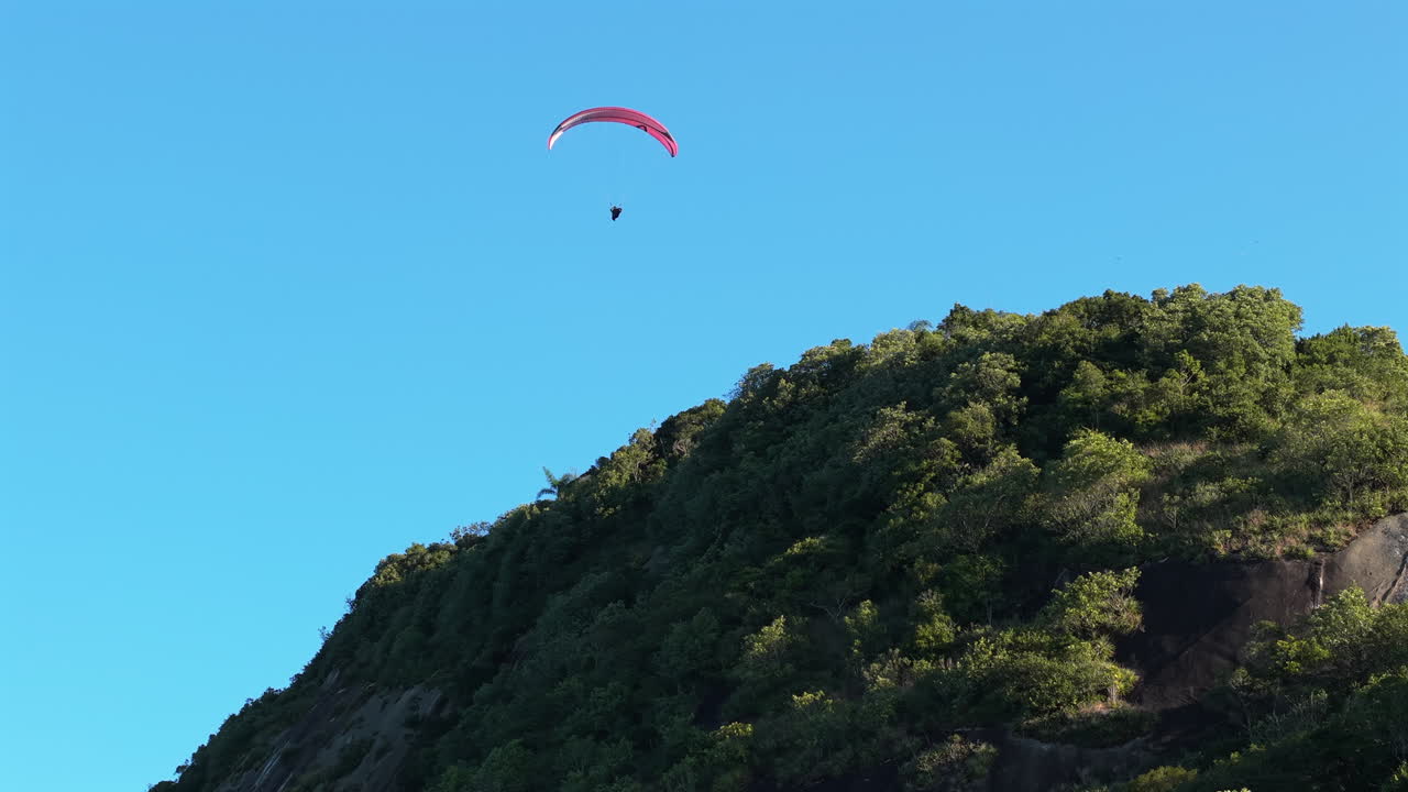 Ground-level upward shot of a red paraglider flying above a lush green hill in Caiobá. The sky is clear blue and the pilot exits the frame