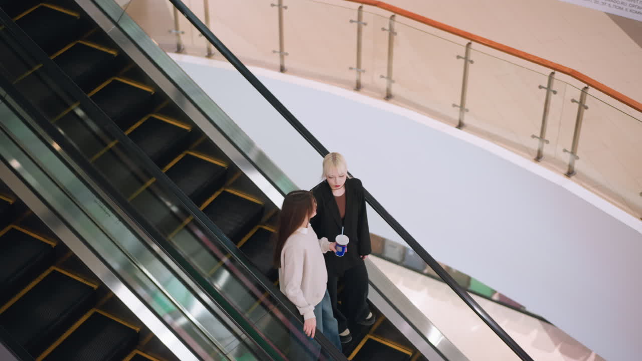 Luftfoto af to kvinder, der går ned ad en rulletrappe i et indkøbscenter og er i en afslappet samtale, mens den ene holder en drink, hvilket skaber en afslappet livsstilsstemning i et moderne indendørs miljø.