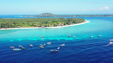 tropical paradise island of white sand filled with green trees surrounded by bright blue sea waters with boats adrift, indonesia - aerial drone shot