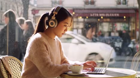 a young woman working at a café: a tranquil scene of focus and creativity with a coffee cup and a laptop, enjoying the atmosphere with headphones on and city life in view