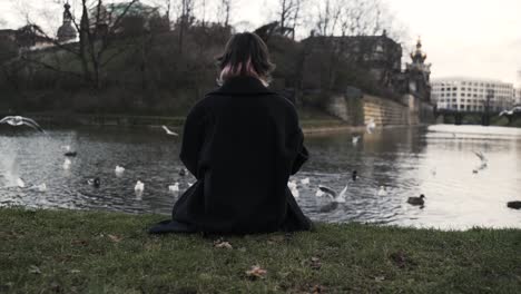 young woman in a black coat sitting in a park in autumn in afternoon and feeding ducks in a sea