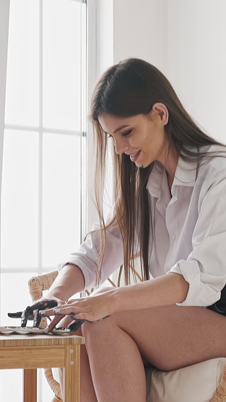 Emotional brunette lady art student in white shirt takes paints by fingers to draw picture on wooden easel by window in studio slow motion side view