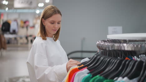 mujer examinando cuidadosamente el estante de ropa, tocando tops vibrantes mientras hace compras en una tienda minorista con fondo borroso de maniquíes y decoración de la tienda