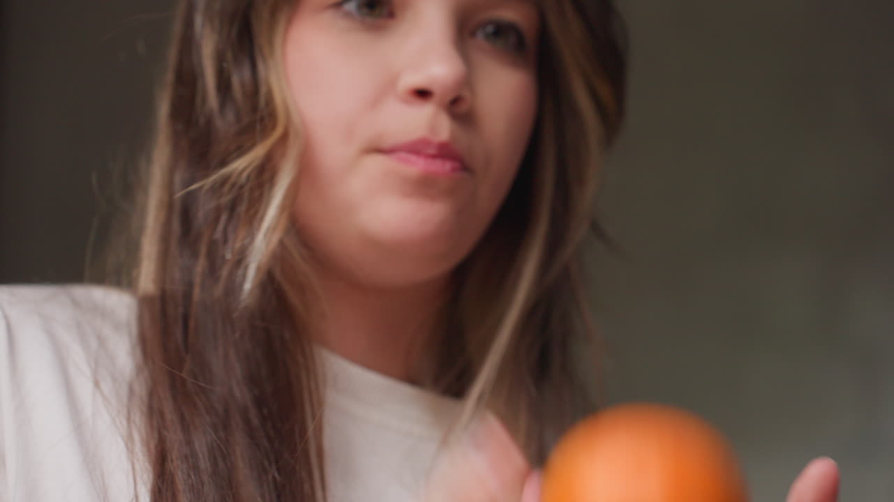 Medium view teenager tossing orange fruit upward then catching with hands, playful motion in indoor light, white sweatshirt visible, soft blur background, relaxed mood, simple daily activity