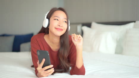 a close-up of a young woman prone on a big comfy bed wearing headphones and enjoying the music from her smartphone