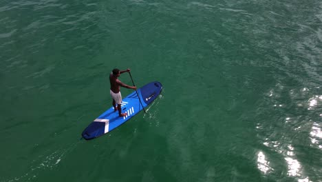 Vista-Aérea-De-Un-Dron-De-Un-Hombre-Asiático-Haciendo-Ejercicio-En-Una-Tabla-De-Remo-En-Aguas-Turquesas-Tropicales-Claras