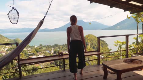 wide dynamic steadicam shot of young woman traveler overlooking volcanic lake of atitlan guatemala from porch of private cabin with hammock