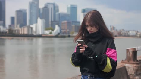 Mittlere-Aufnahme-Eines-Besorgten-Mädchens,-Das-Mittags-In-Der-Nähe-Des-Flusses-Mit-Einer-Stadt-Im-Hintergrund-Auf-Ihrem-Handy-Chattet