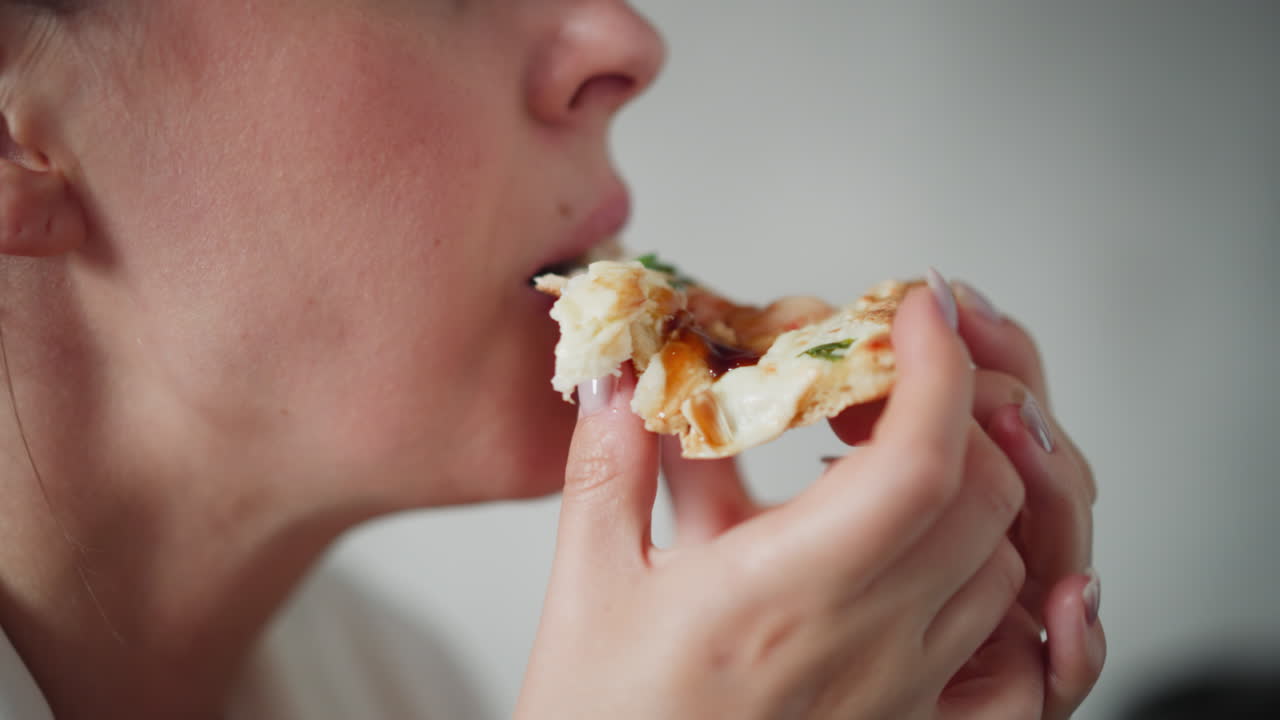 Close up of young girl enjoying cheesy pizza bite with melted toppings and fresh herbs, holding pizza slice near lips, savoring delicious flavor, background softly blurred