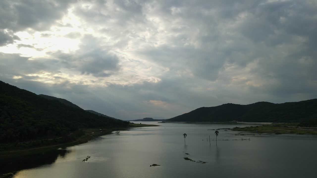 Aerial reverse footage of this fantastic landscape of the lake with rainclouds, sun setting, palm trees in the middle of the lake; Saraburi, Thailand