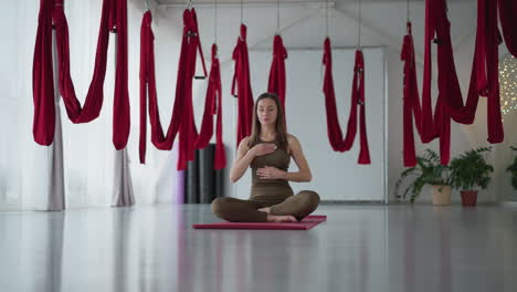young lady sitting cross legged on red mat under silk hammocks in bright studio space practicing mindful yoga with eyes closed and gentle breathing focusing on inner peace serenity