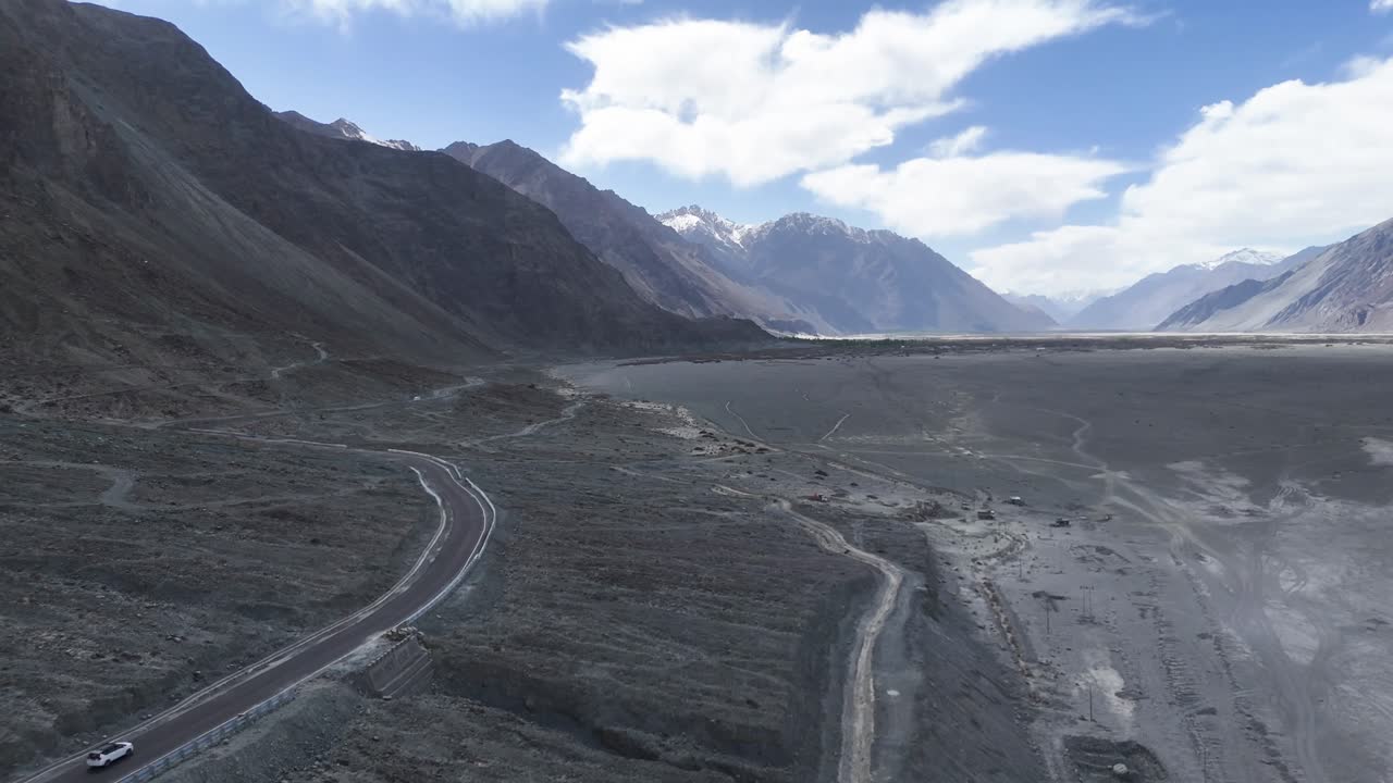Aerial drone shot following a lone vehicle traversing the remote roads of Nubra Valley, highlighting the valley’s vastness.