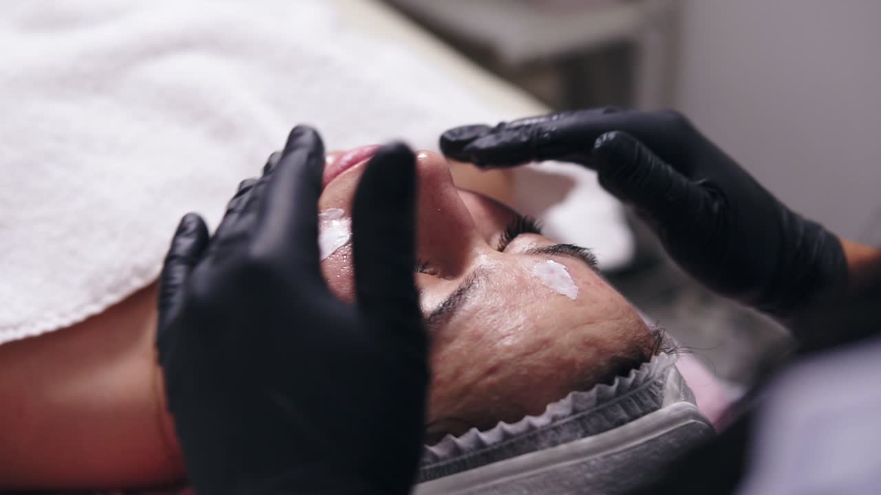 Close Up view of a woman with blemish skin lying on the couch while professional cosmetologist is applying face cream and making