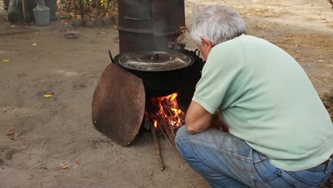 man puts branches in the fire while cooking fish stew in a pot outdoors