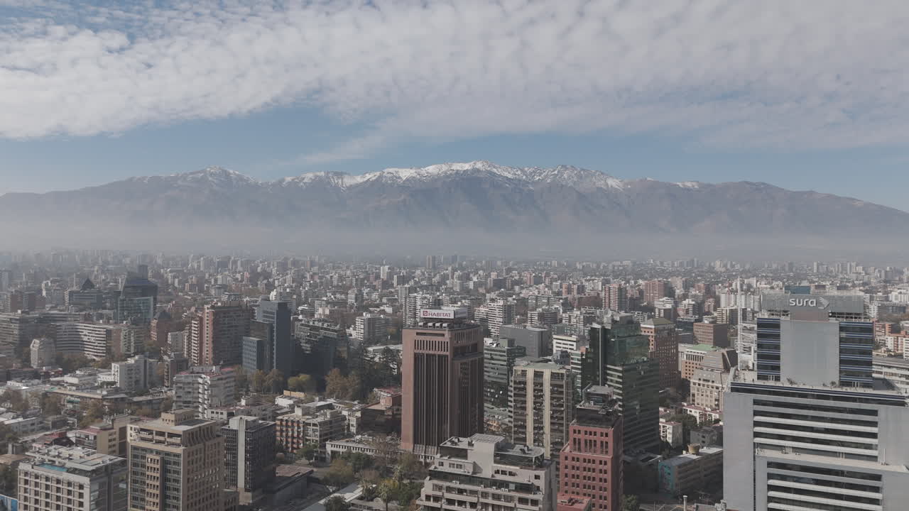 Backwards drone shot of Santiago de Chile in South America watching over the skyscrapers and big buildings with snowy mountains in the background on bright but cloudy day LOG
