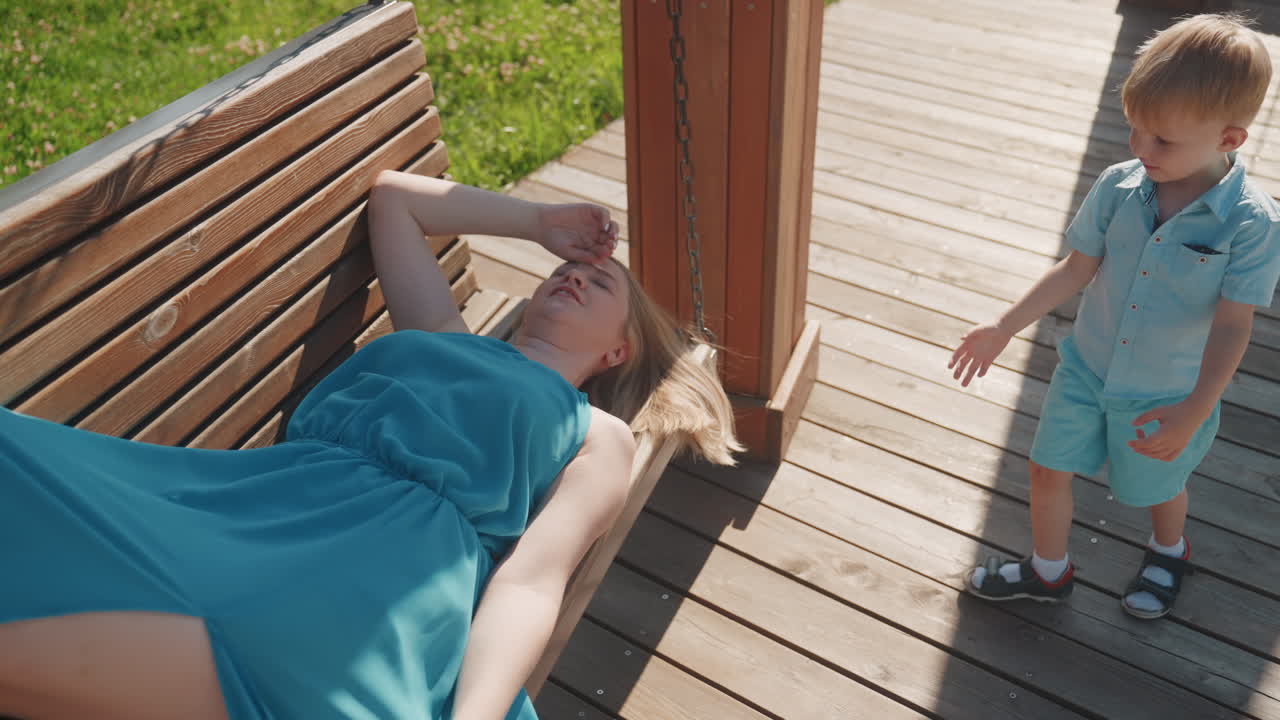 Caucasian child leans over resting mother on swing bench in park weekend little boy touches mothers hand and checks comfort, sunlit deck boards, intimate family interaction, caring moment