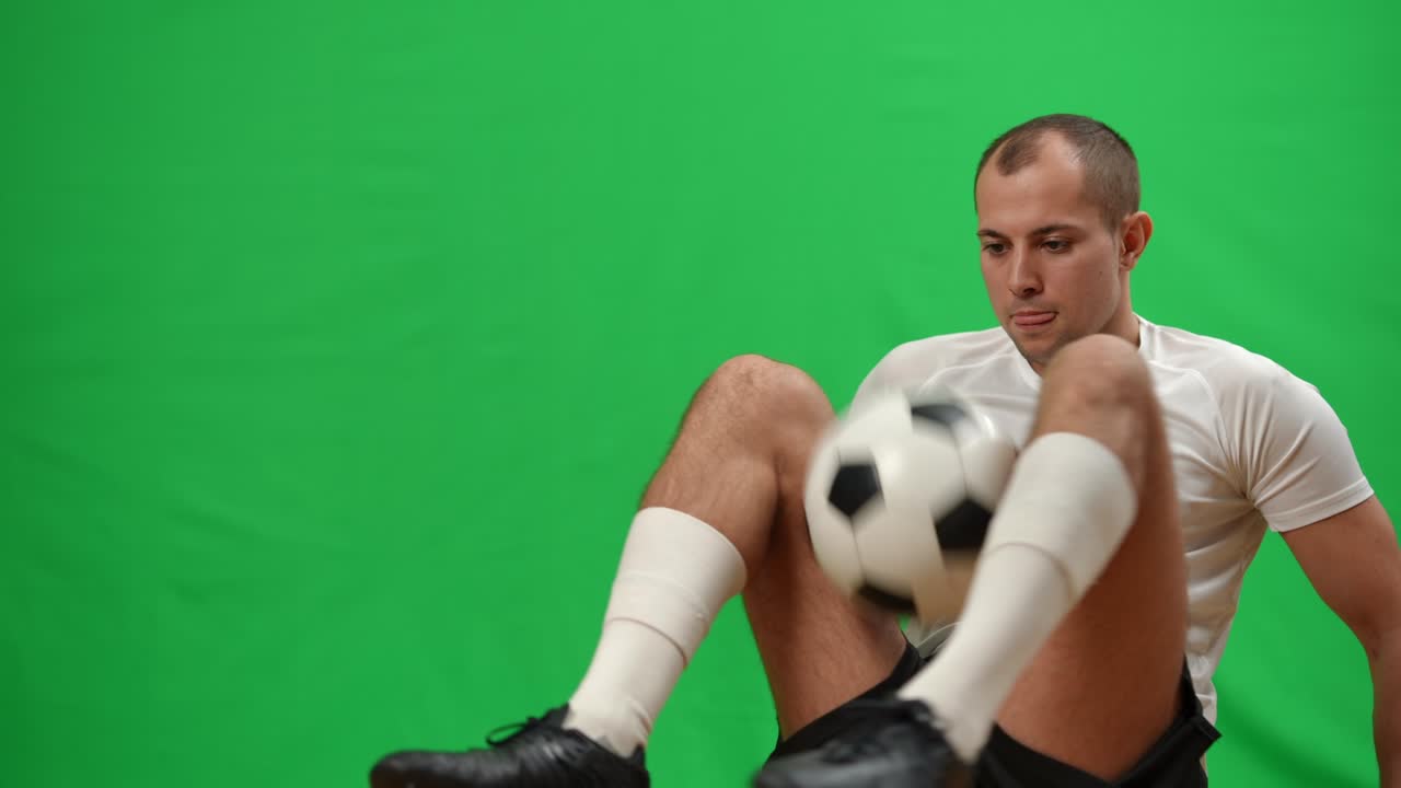 프로 축구 선수가 녹색 스크린에 앉아서 웃으며 멀리 바라보는 다리로 공을 잡고 글링을 하고 있다. 긍정적 인 백인 스포츠 선수가 크로마키 배경에서 운동하고 있다.