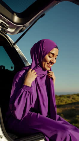 a young woman in a purple hijab smiling outdoors next to a car