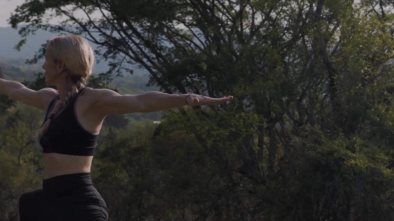 Woman holds a calm warrior pose with arms outstretched above a wooded hillside