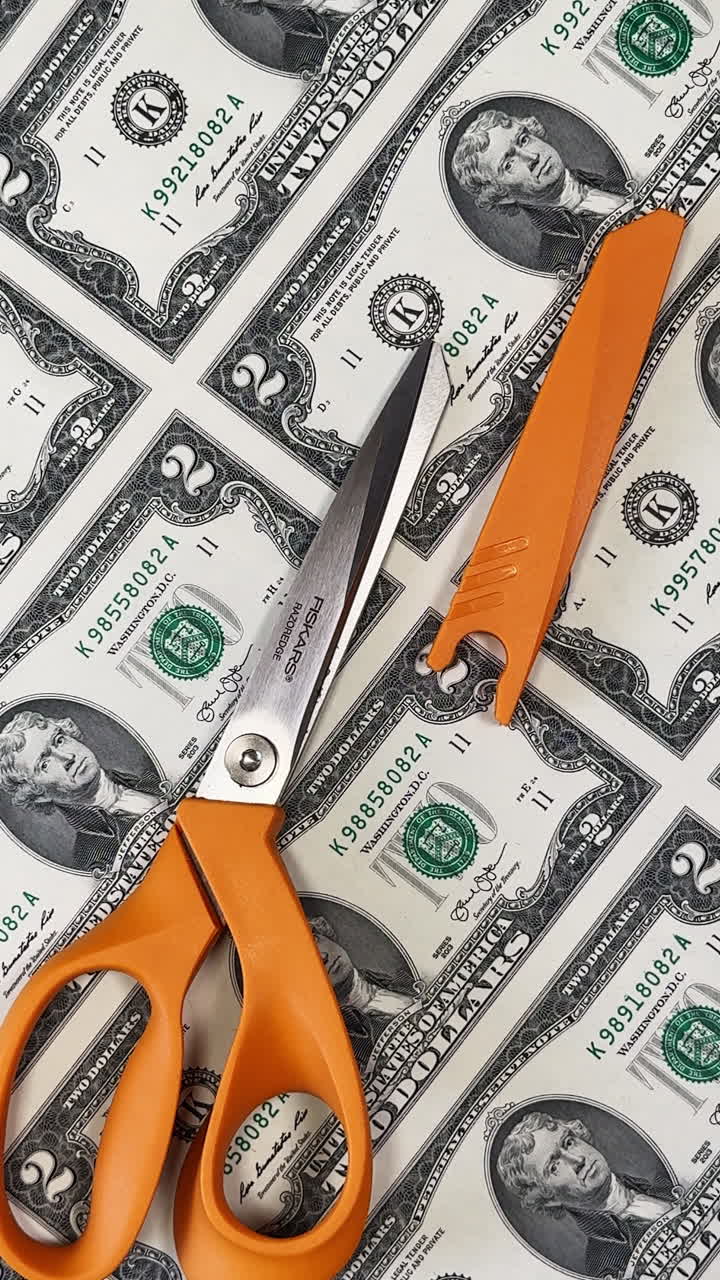 San Antonio, USA, 14 August 2025: Big scissors with orange handles lie on the sheet of uncut two dollar banknotes. Money printing concept. Close up. Vertical video