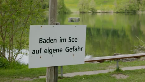 see im freien mit badeplattform in grüner landschaft mit schild in deutscher sprache, deutscher text übersetzung: schwimmen im see auf eigene gefahr am see, fokus stark auf das textfeld, kamera panning schild zu ring 4k uhd prores hq 10bit 25p