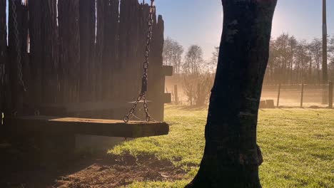 a wood swing floats above the grass near a tree and wood fence on a foggy morning, medium shot