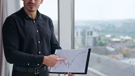 male hand following the graph printed on the paper. businessman in black clothes pointing at chart and talking. office indoor backdrop.